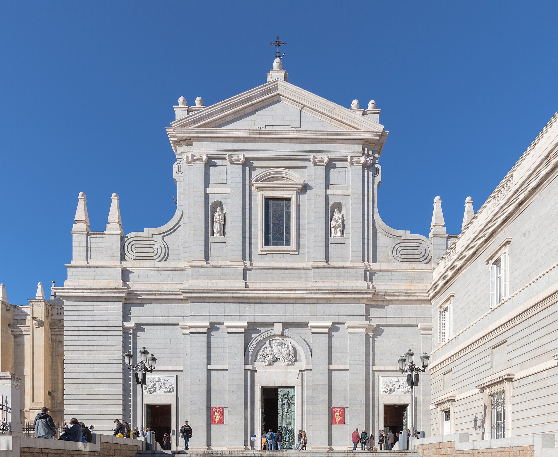 Fachada principal de la catedral de la Almudena en Madrid: frente de piedra blanca con frontón triangular coronado por una cruz, dos estatuas en nichos flanqueando el cuerpo central y una portada de arco adovelado bajo el relieve, con varias personas en la plaza.