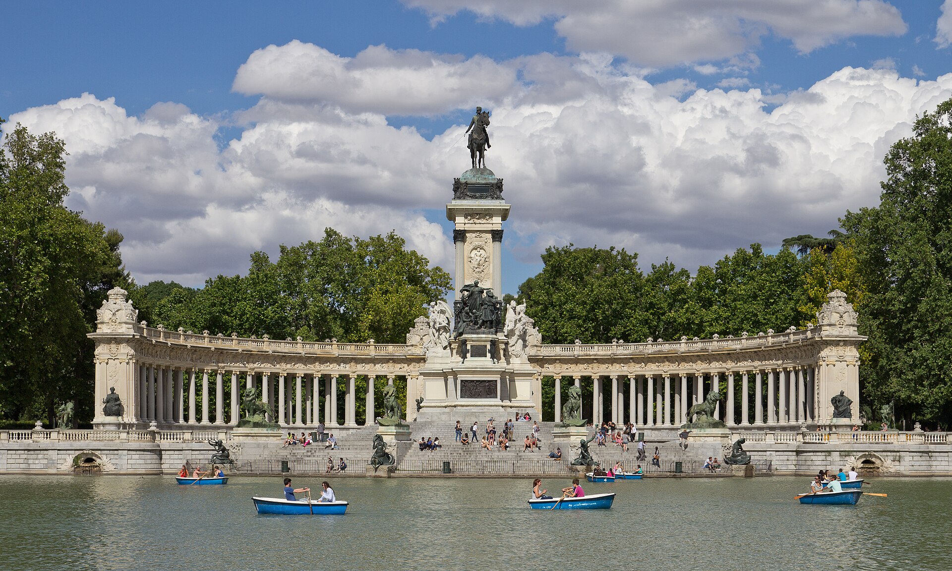 Monumento a Alfonso XII en los Jardines del Retiro visto desde el estanque: columnata semicircular de mármol con un alto pedestal central coronado por una estatua ecuestre, y barcas de remo sobre el agua delante.