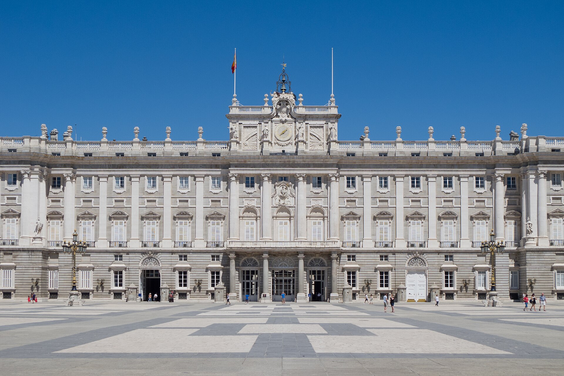 Fachada sur del Palacio Real de Madrid vista desde la plaza de la Armería: tres plantas de piedra con el reloj central bajo un templete, la bandera de España en la balastrada y la plaza empedrada delante con visitantes.